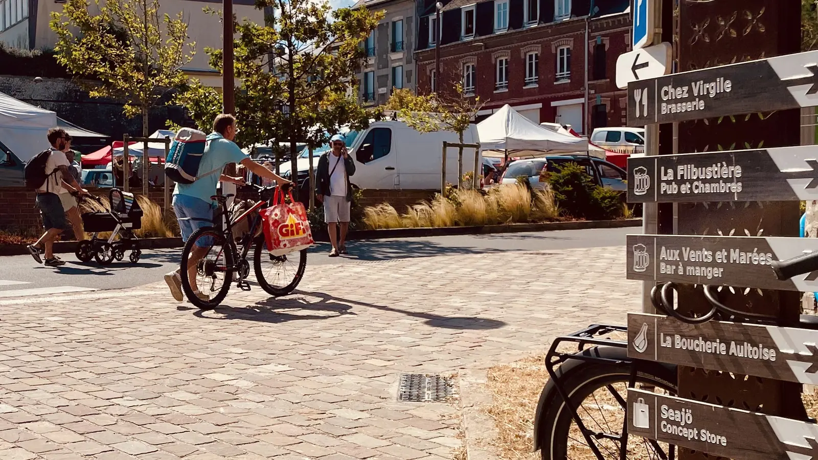 Cyclistes sur la place d'Ault, devant les panneaux qui indiquent les commerces